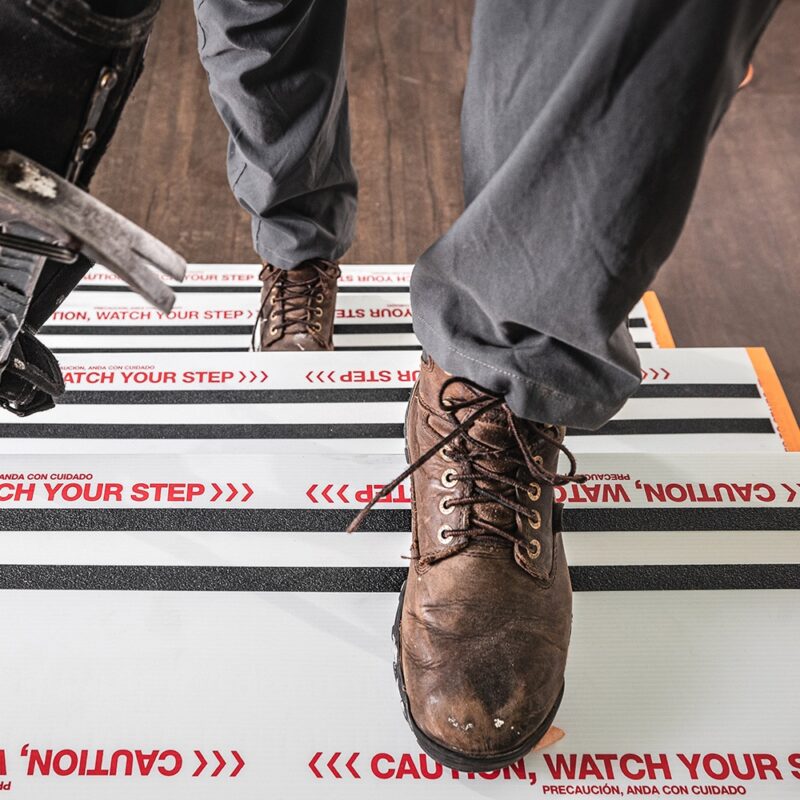 A person wearing brown boots steps on a Stair Shield striped floor mat printed with "CAUTION, WATCH YOUR STEP" in red, positioned beside metal equipment.
