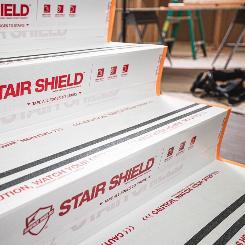 Stair Shield white stair protection covers are taped to indoor stairs at a construction site, with tools and equipment in the background.