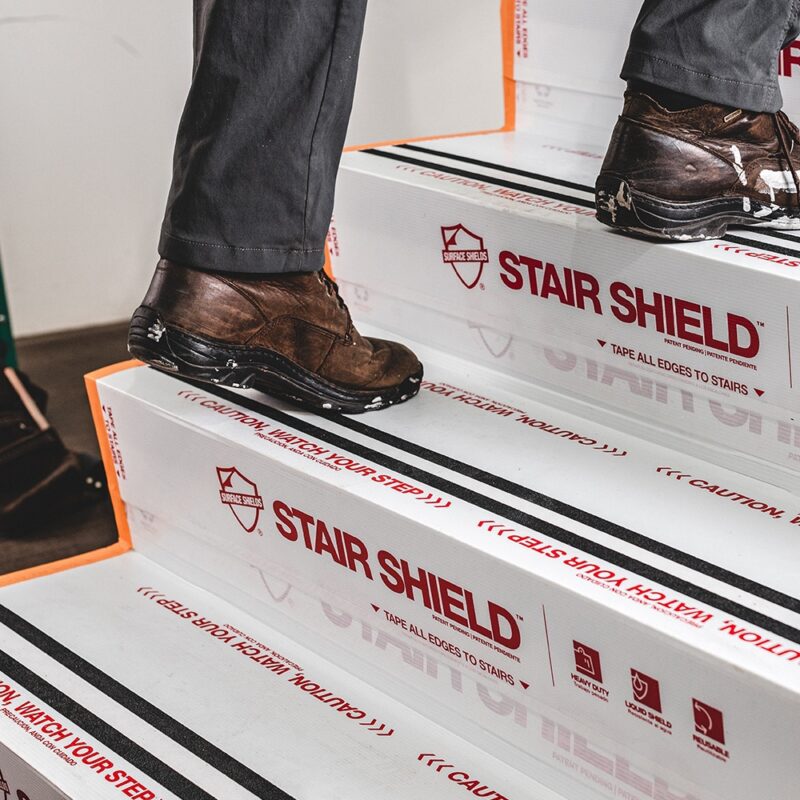 Two people walk on stairs covered with Stair Shield, a protective material labeled with caution signs and "WATCH YOUR STEP" warnings.